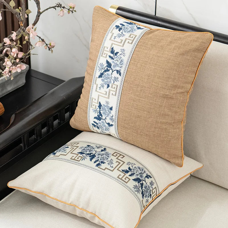 Deux coussins décoratifs en tissu beige et motifs bleus et gris ornent un canapé, accompagnés d'une branche fleurie dans un vase.