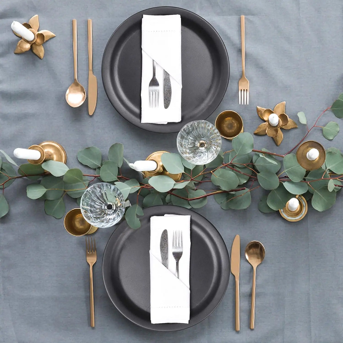 Élégante table de fête avec des assiettes noires, des couverts dorés, des bougies et une décoration végétale.