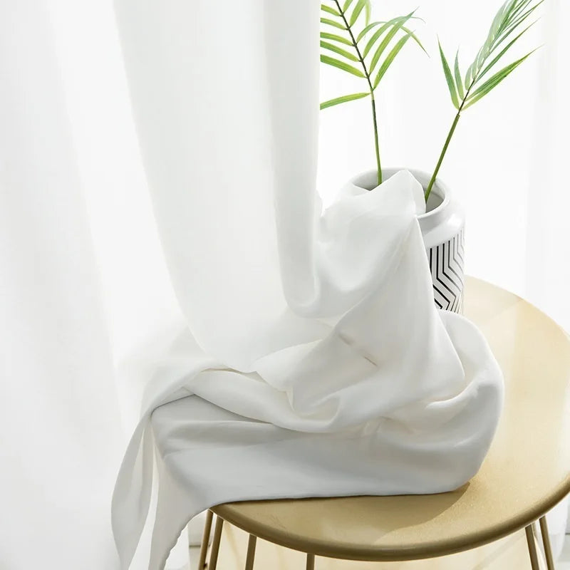 Rideau blanc élégant tombant gracieusement sur une table, accentuée par un vase décoratif moderne avec des plantes vertes. Éclairage naturel doux offrant une ambiance lumineuse et apaisante pour tout intérieur. Idéal pour ajouter une touche de sophistication à votre décoration.