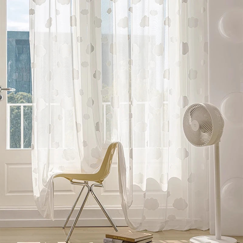 Voilage blanc transparent à motifs de nuages, flottant doucement devant une baie vitrée, avec une chaise design et un ventilateur sur pied.