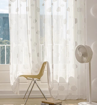 Voilage blanc transparent à motifs de nuages, flottant doucement devant une baie vitrée, avec une chaise design et un ventilateur sur pied.