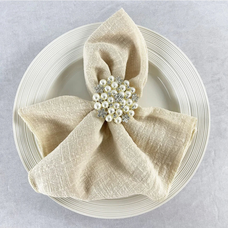 Un arrangement raffiné de serviette beige avec une broche ornée de perles nacrées et de cristaux brillants, placé sur une assiette blanche texturée, créant une ambiance élégante et sophistiquée.