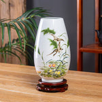 Un vase décoratif en porcelaine blanche avec une scène colorée d'une végétation luxuriante et d'un oiseau volant, posé sur un socle en bois.
