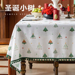 Nappe de table élégante et festive aux motifs d'arbres de Noël verts et dorés, ornée de pompons et d'autres décorations de Noël sur une table décorée pour les fêtes.