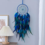Un magnifique attrape-rêves bleu et vert orné de plumes de couleurs vibrantes, suspendu sur un mur blanc, avec une lampe en arrière-plan.