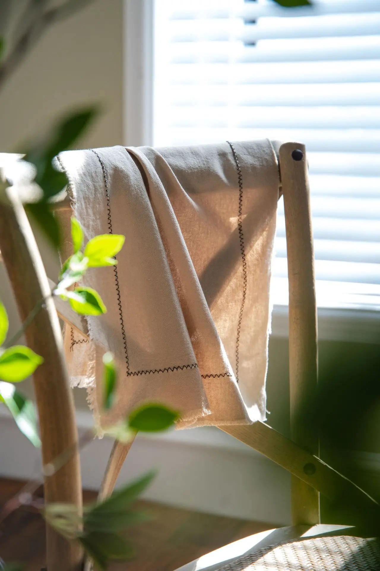 Une couverture en lin beige avec des détails cousus, posée sur une chaise en bois près d'une fenêtre avec des plantes vertes.