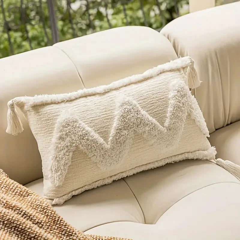 Coussin décoratif avec motifs triangulaires et franges texturées, posé sur un canapé beige dans un environnement naturel.