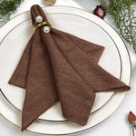 Pliage de serviette de table en lin brun orné de perles, sur un plat blanc avec des décors floraux.