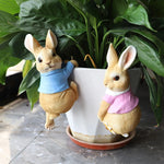 Deux lapins en peluche dans un pot de fleurs, l'un vêtu d'un pull bleu et l'autre d'un pull rose, entourés de feuilles vertes.