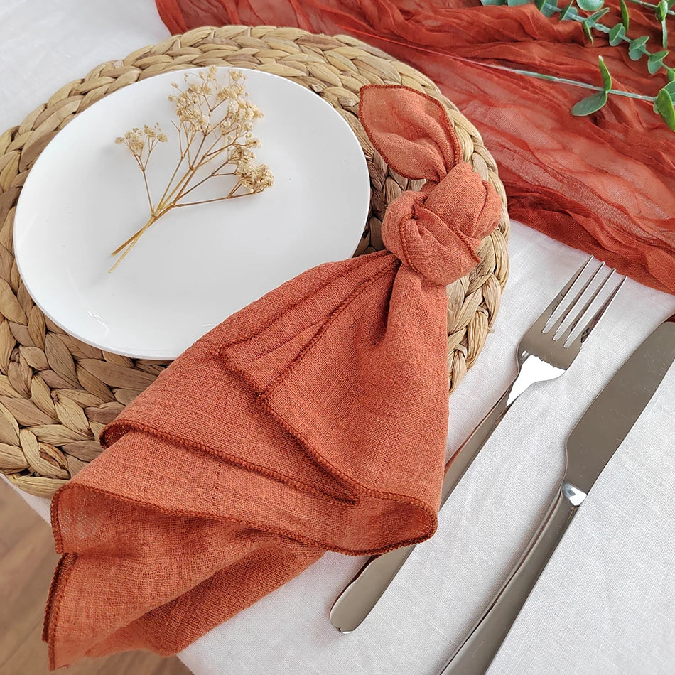 Une assiette blanche avec une tige de fleurs séchées posée dessus, entourée d'un napperon tressé et d'un linge de table de couleur terracotta, avec des couverts disposés à côté.