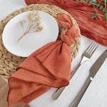 Une assiette blanche avec une tige de fleurs séchées posée dessus, entourée d'un napperon tressé et d'un linge de table de couleur terracotta, avec des couverts disposés à côté.