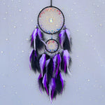 Un capteur de rêves décoratif violet et noir, fait main avec des plumes et des perles colorées disposées dans un motif de toile d'araignée, suspendu devant un ciel étoilé.