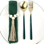 Une nappe vert foncé avec des perles de bois blanc et un gland décoratifs, accompagnée de couverts en métal doré sur une assiette blanche.