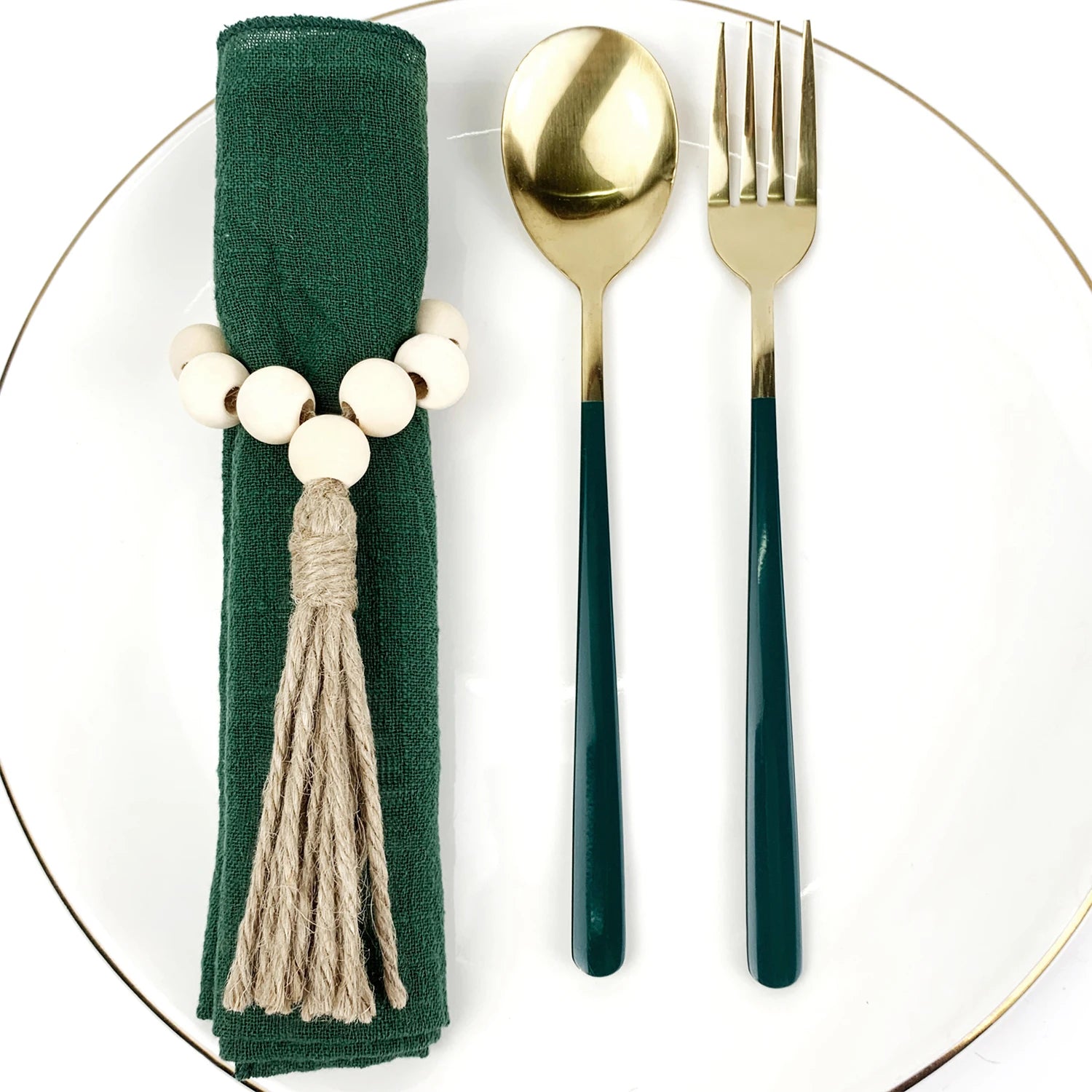 Une nappe vert foncé avec des perles de bois blanc et un gland décoratifs, accompagnée de couverts en métal doré sur une assiette blanche.