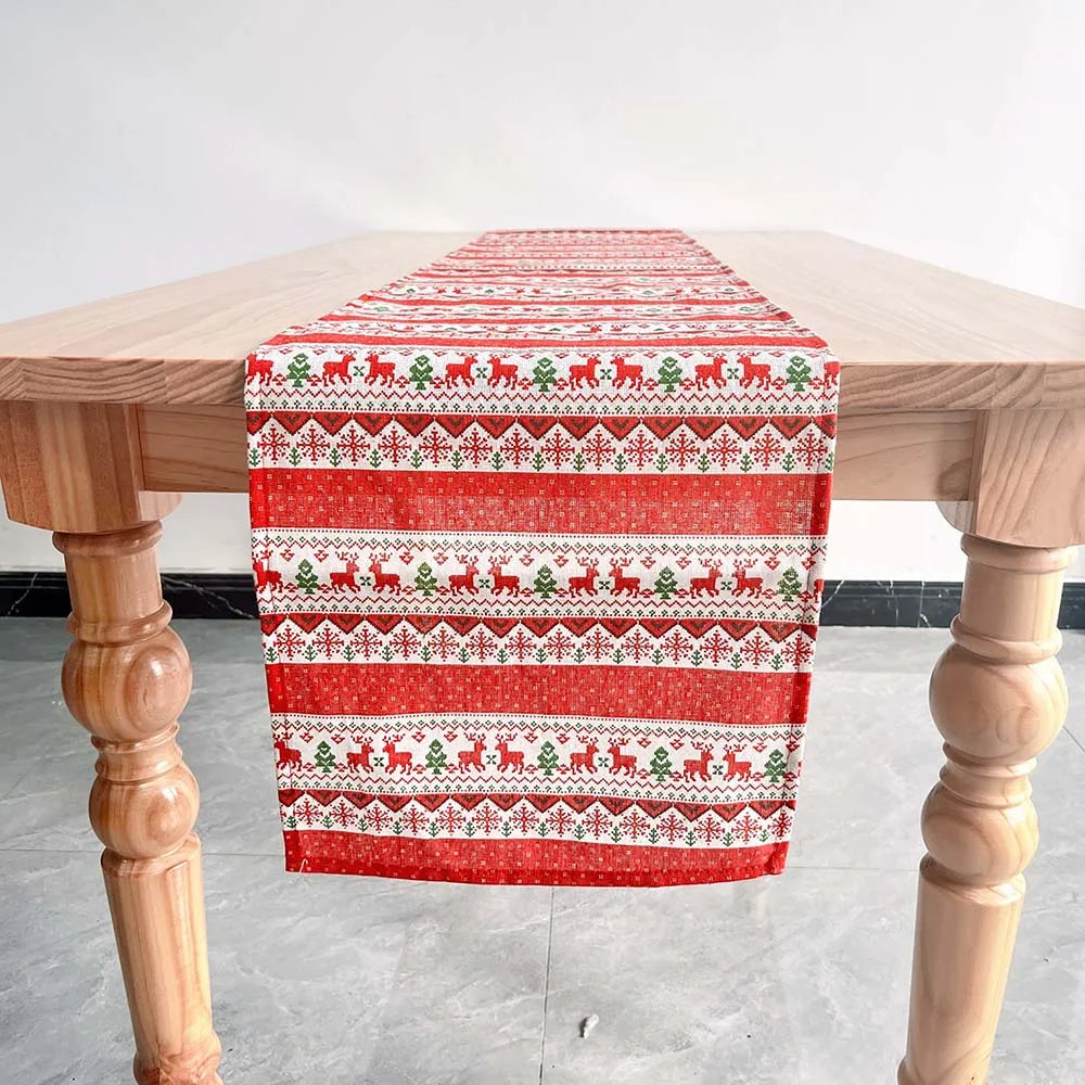 Une nappe de table en coton avec des motifs de rennes et de flocons de neige dans des tons rouges et blancs, posée sur une table en bois.