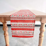 Une nappe de table en coton avec des motifs de rennes et de flocons de neige dans des tons rouges et blancs, posée sur une table en bois.