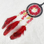 Un capteur de rêves décoratif avec un anneau noir et des plumes rouges, décoré de pierres précieuses, pendant sur un fond blanc.