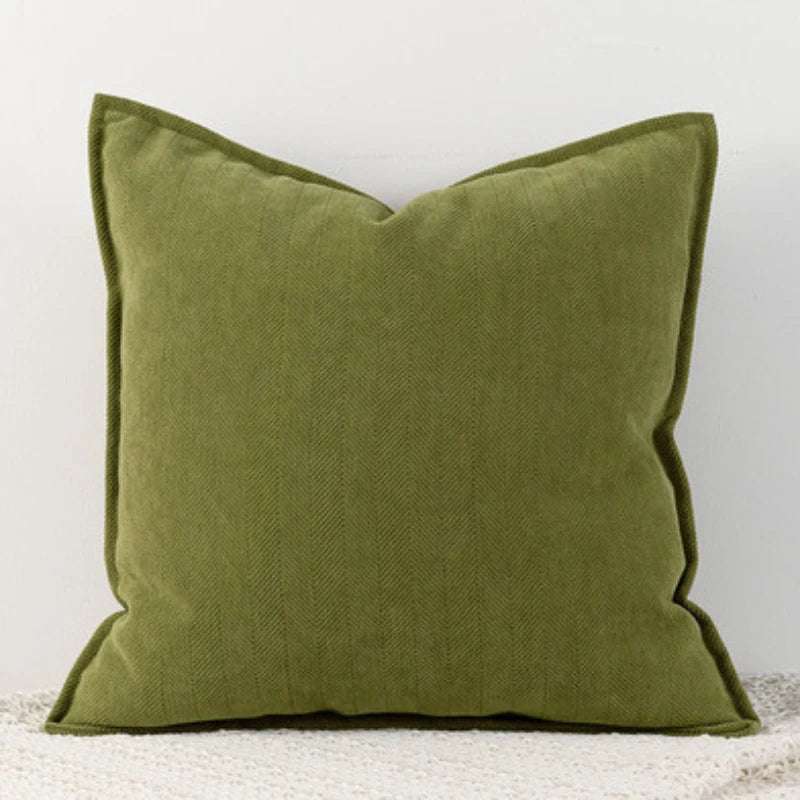 Coussin décoratif carré en tissu vert olive aux motifs texturés, posé sur un sol gris.