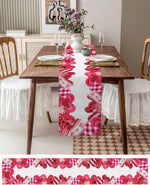 Une table de salle à manger élégamment dressée avec un chemin de table à motifs rouges, des fleurs dans un vase blanc et des couverts sur des assiettes blanches.