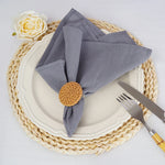 L'image montre une assiette blanche sur un dessous de plat en tressage beige, avec une serviette grise pliée et retenue par un support en osier doré, ainsi qu'une rose blanche sur le côté.