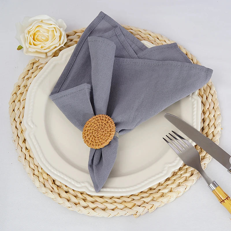 L'image montre une assiette blanche sur un dessous de plat en tressage beige, avec une serviette grise pliée et retenue par un support en osier doré, ainsi qu'une rose blanche sur le côté.