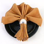 Un arrangement décoratif de serviettes en tissu ocre, roulées et attachées avec un nœud argenté, sur un plat noir avec des reflets scintillants.