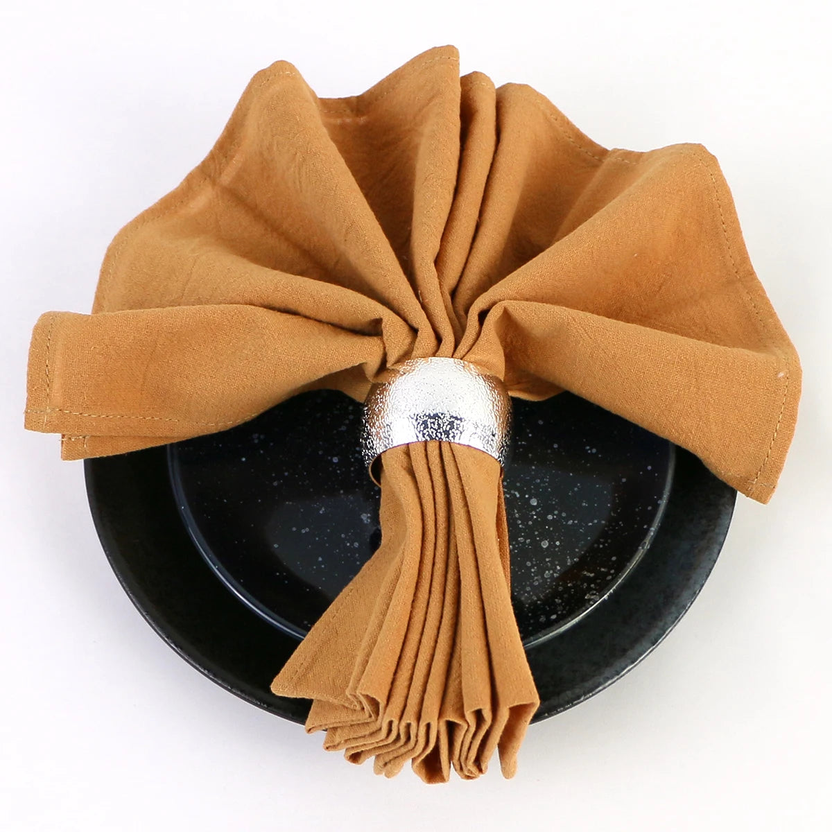 Un arrangement décoratif de serviettes en tissu ocre, roulées et attachées avec un nœud argenté, sur un plat noir avec des reflets scintillants.