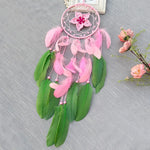 Un capteur de rêves rose et vert avec des plumes et une fleur décorative suspendu devant un fond gris et décoré de fleurs en fleurs.