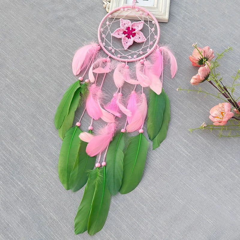 Un capteur de rêves rose et vert avec des plumes et une fleur décorative suspendu devant un fond gris et décoré de fleurs en fleurs.