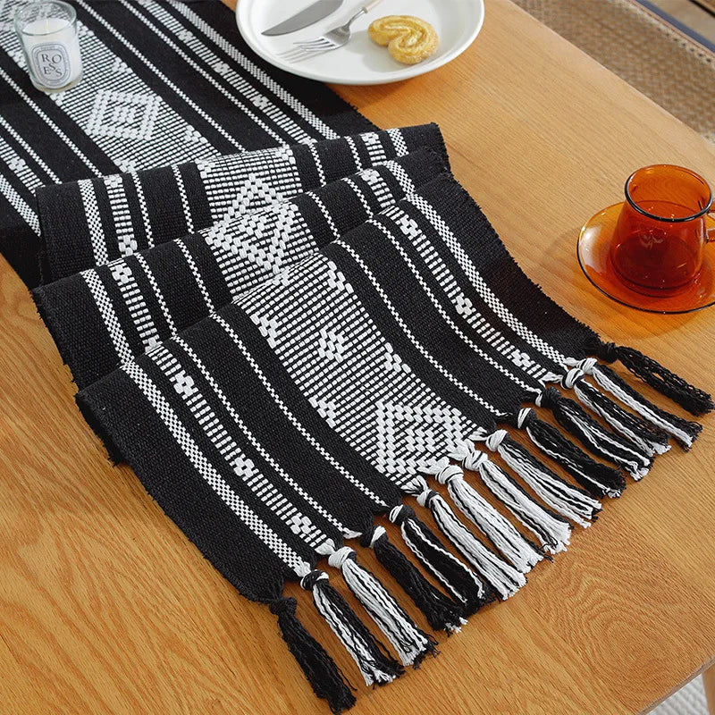 Sur une table en bois, on peut voir un plat recouvert d'un motif géométrique noir et blanc à franges, avec une tasse en verre à côté.