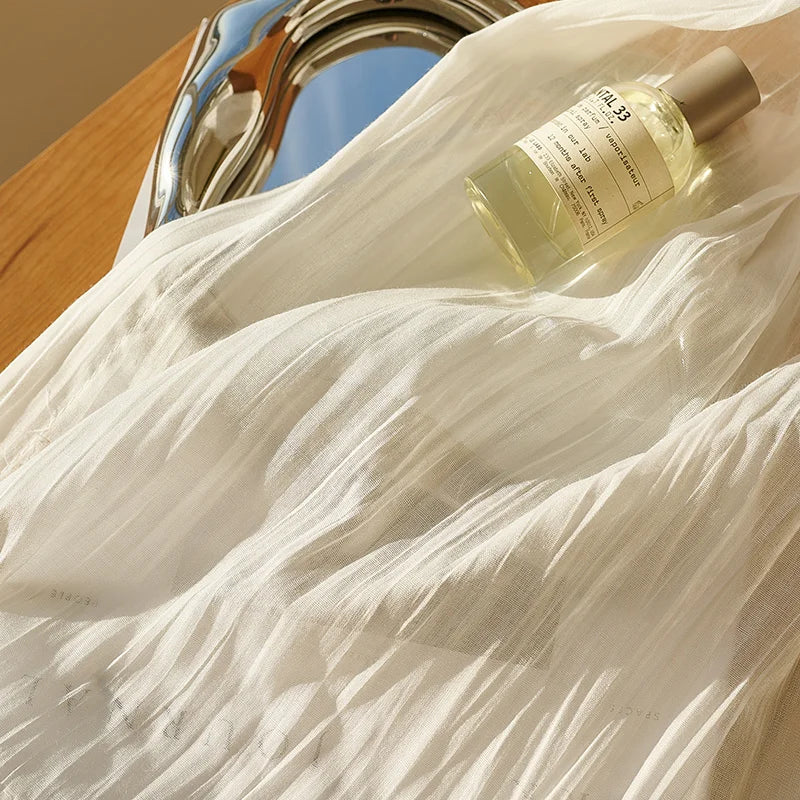 Détail d’un voilage d’été plissé avec flacon de parfum et plateau chromé, capturant une lumière dorée et estivale.
