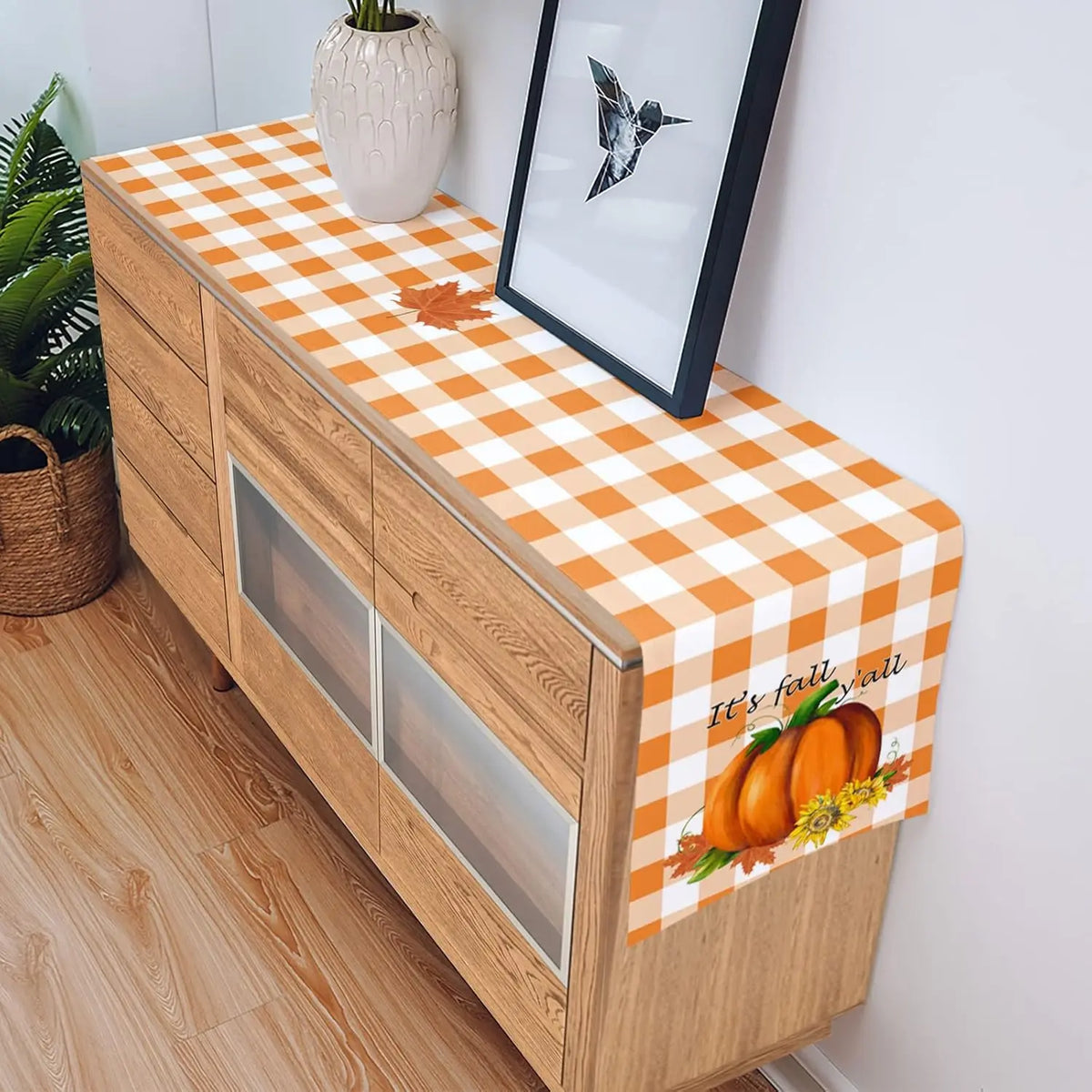 Élégant buffet en bois avec une nappe à carreaux oranges et blancs, décoré d'une feuille d'érable et d'un photophore en forme de citrouille, reflétant l'esprit de l'automne.