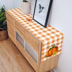 Élégant buffet en bois avec une nappe à carreaux oranges et blancs, décoré d'une feuille d'érable et d'un photophore en forme de citrouille, reflétant l'esprit de l'automne.