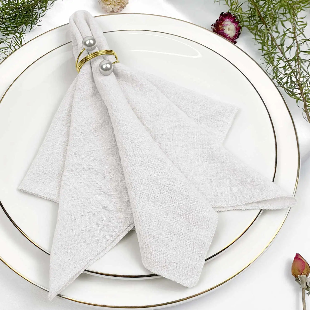 Serviette blanche élégamment disposée sur une assiette blanche avec des anneaux dorés et des fleurs décoratives.