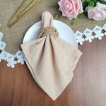 Serviette de table beige nouée avec une corde, placée sur une nappe en jute, entourée de roses roses, de feuilles vertes et de petites fleurs blanches.