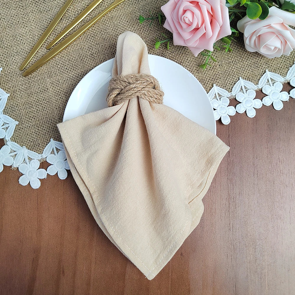 Serviette de table beige nouée avec une corde, placée sur une nappe en jute, entourée de roses roses, de feuilles vertes et de petites fleurs blanches.