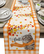 Une décoration de table d'automne chaleureuse avec un motif de feuilles multicolores et un chat portant une écharpe orange, sur un chemin de table avec des carreaux.