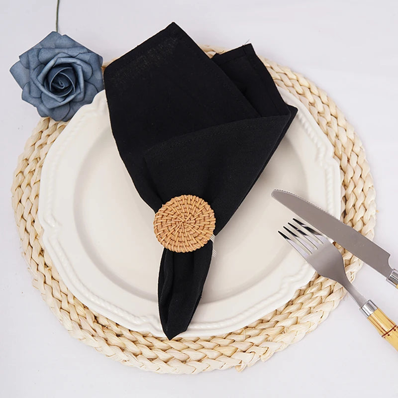 Une nappe ronde en jonc de mer beige accueille une assiette blanche sur laquelle sont posés un set de table noir, un élément décoratif en rotin et une rose bleu pâle, créant une composition élégante et épurée.