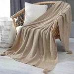 Couverture douillette beige en tricot, avec des motifs texturés, posée sur un fauteuil en rotin, créant une ambiance chaleureuse et cocooning dans l'intérieur.