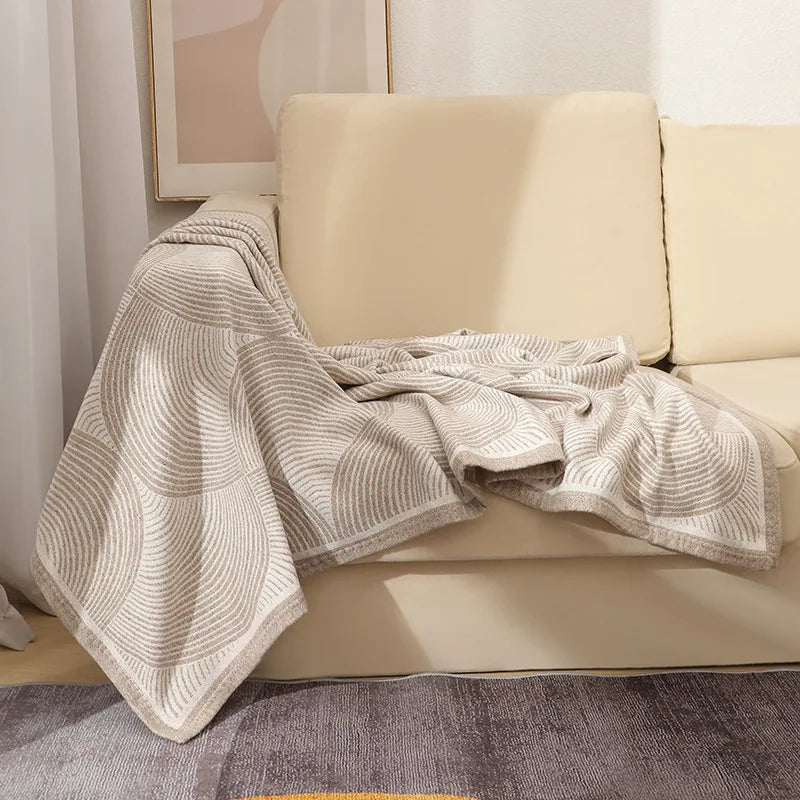 Une couverture douillette en tricot gris clair ornée de motifs rayés repose sur un canapé beige dans un intérieur chaleureux et confortable.
