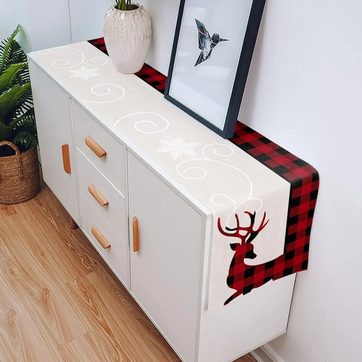 Meuble blanc moderne avec motifs décoratifs, plateau en bois, décoration florale et photo encadrée sur un fond de tissu à carreaux rouges et noirs.