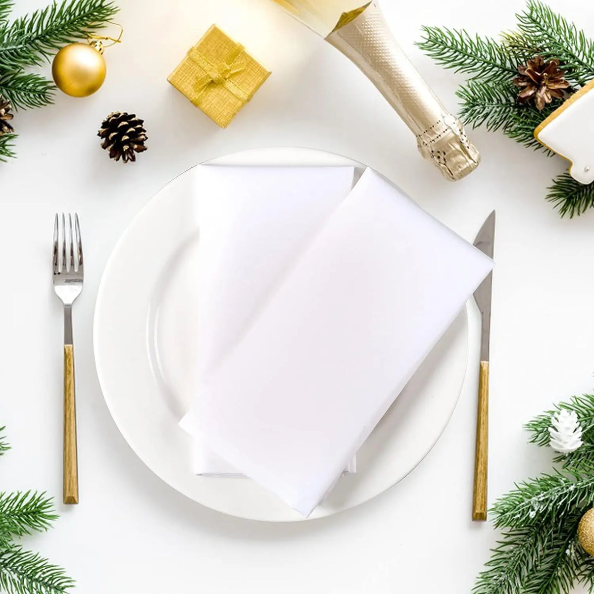 La table de Noël blanche et élégante est décorée de branches de sapin vertes, de pommes de pin, d'un cadeau doré, et comprend une assiette, une fourchette et un couteau dorés, créant une ambiance festive et chaleureuse.