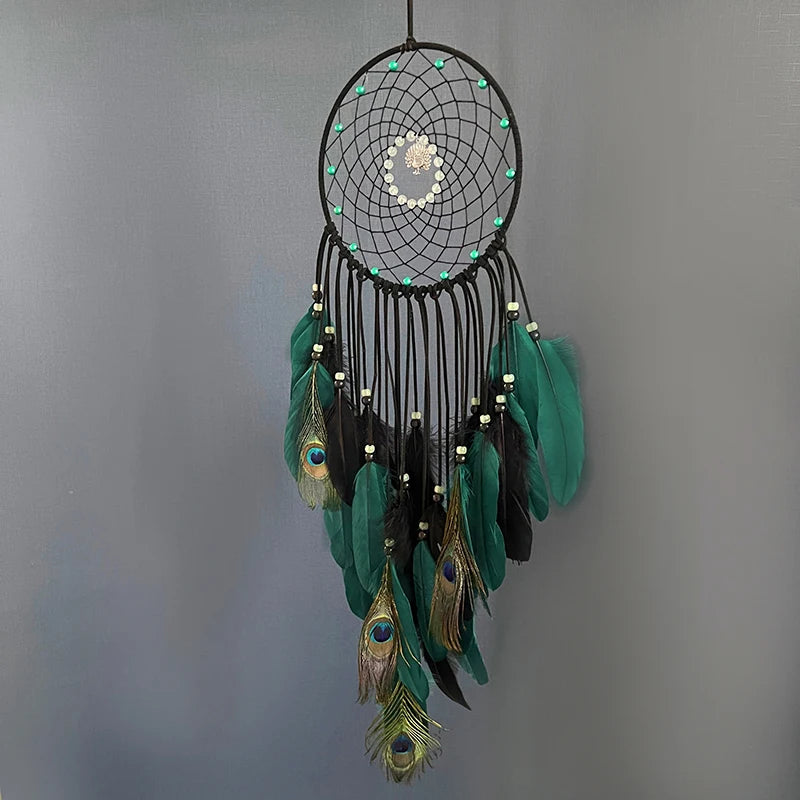 Un capteur de rêves circulaire aux plumes vertes, bleu paon et perles blanches, suspendu à l'aide de cordes noires.