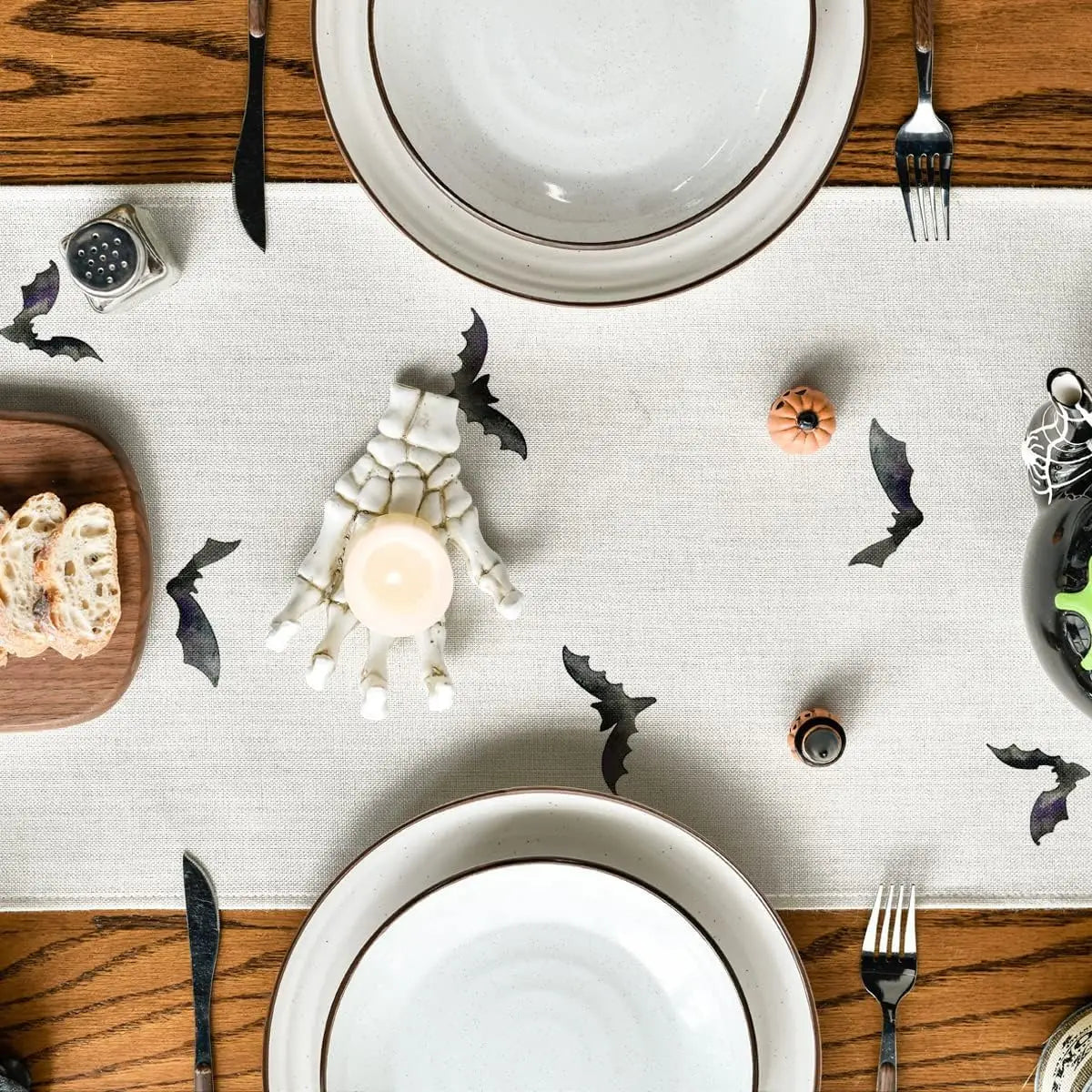 La table de salle à manger est joliment décorée pour l'Halloween, avec des assiettes blanches, des bougies et des chauves-souris en papier noirs disposés sur un fond en bois et un chemin de table clair.
