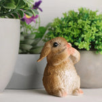 Une figurine mignonne d'un lapin en céramique, assis dans un environnement naturel avec des plantes verdoyantes en arrière-plan.
