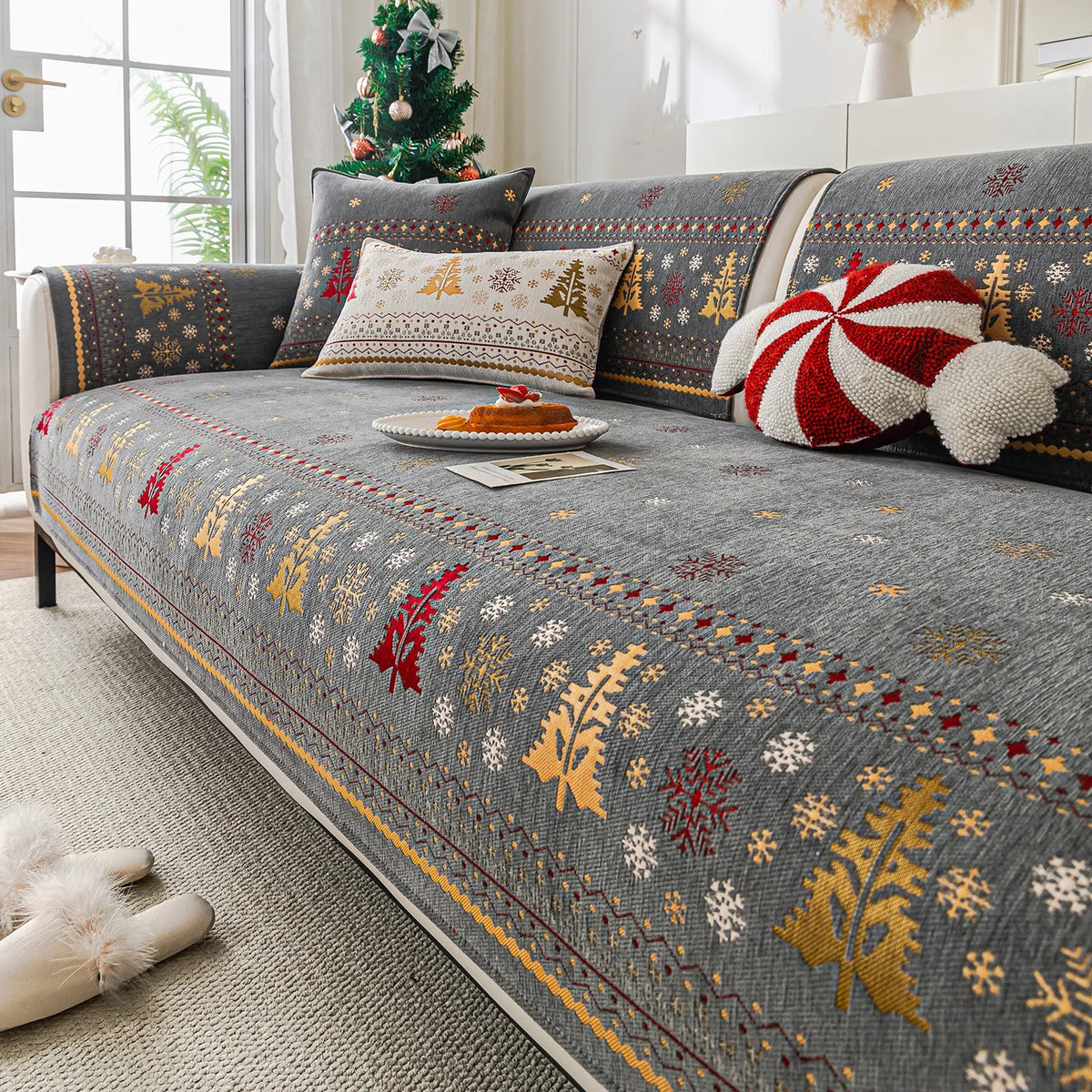 Un canapé douillet décoré de motifs d'hiver colorés sur un tissu gris, accompagné de coussins décoratifs et d'un sapin de Noël dans le fond, créant une ambiance chaleureuse et festive.