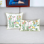 Deux coussins décoratifs aux motifs floraux colorés et riches en détails, posés sur un canapé gris, avec une œuvre d'art encadrée au mur en arrière-plan.