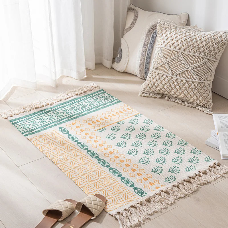 Un tapis décoratif avec des motifs géométriques colorés et une finition à franges, placé dans un intérieur lumineux avec des coussins de style bohème.