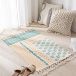 Un tapis décoratif avec des motifs géométriques colorés et une finition à franges, placé dans un intérieur lumineux avec des coussins de style bohème.