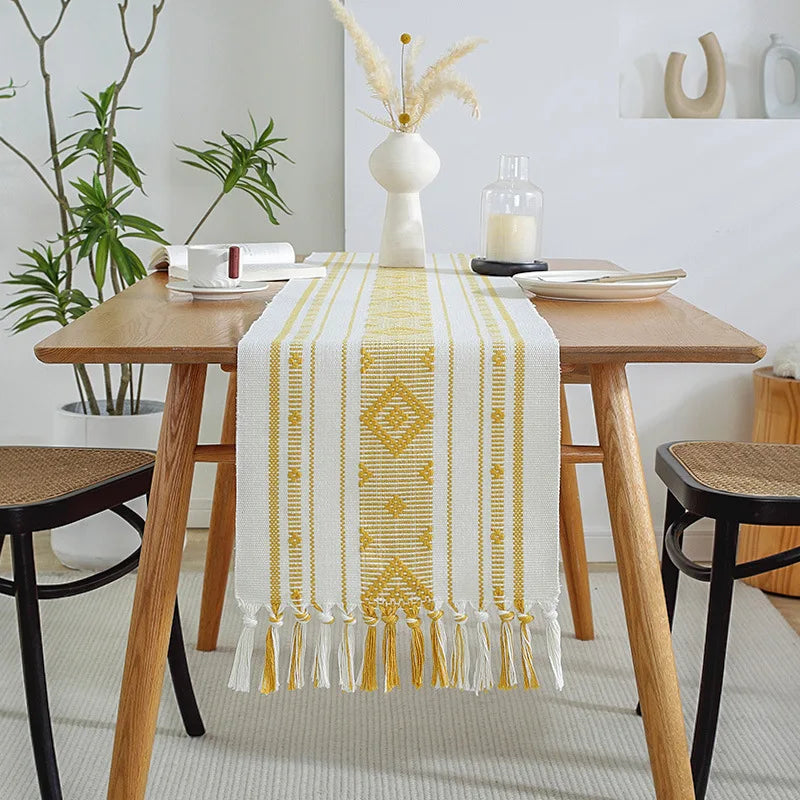 Une table en bois moderne avec un chemin de table orné de motifs géométriques jaunes et de franges blanches, dans un décor intérieur épuré et élégant.
